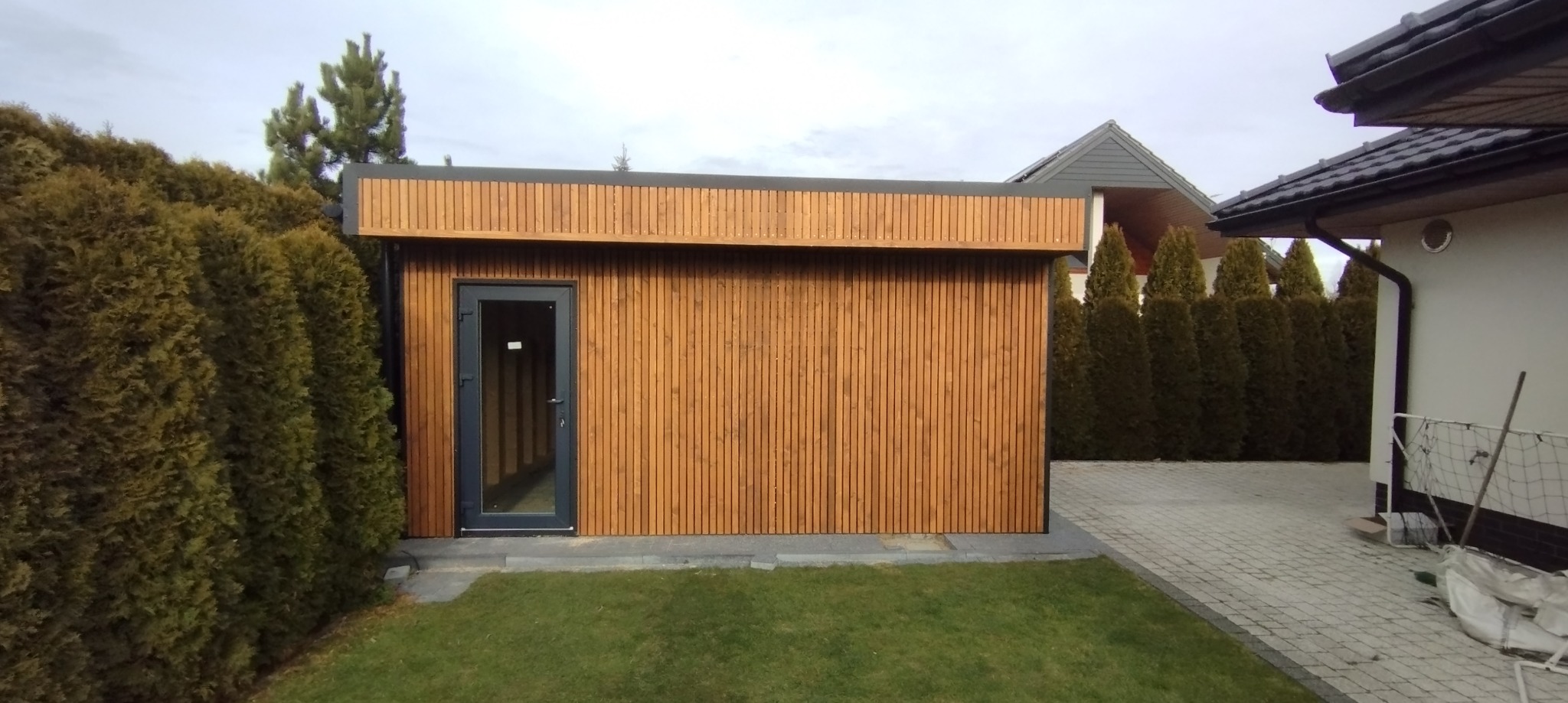 Nowoczesna, drewniana konstrukcja z ciemną stolarką drzwiową, otoczona zielenią. Widoczne pionowe deski elewacyjne i starannie wykończone detale.