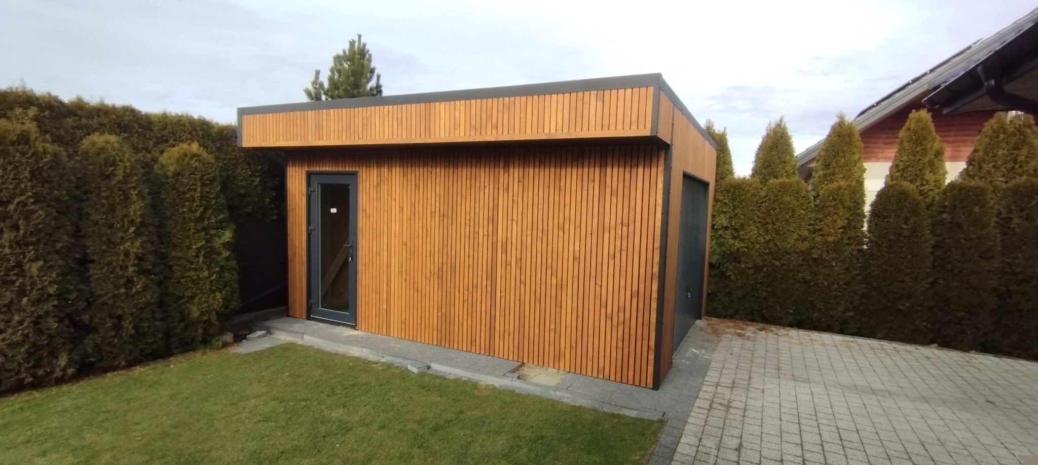 Nowoczesna, drewniana konstrukcja z drzwiami i bramą garażową, otoczona zielenią. Elewacja z pionowych desek w ciepłym odcieniu brązu.