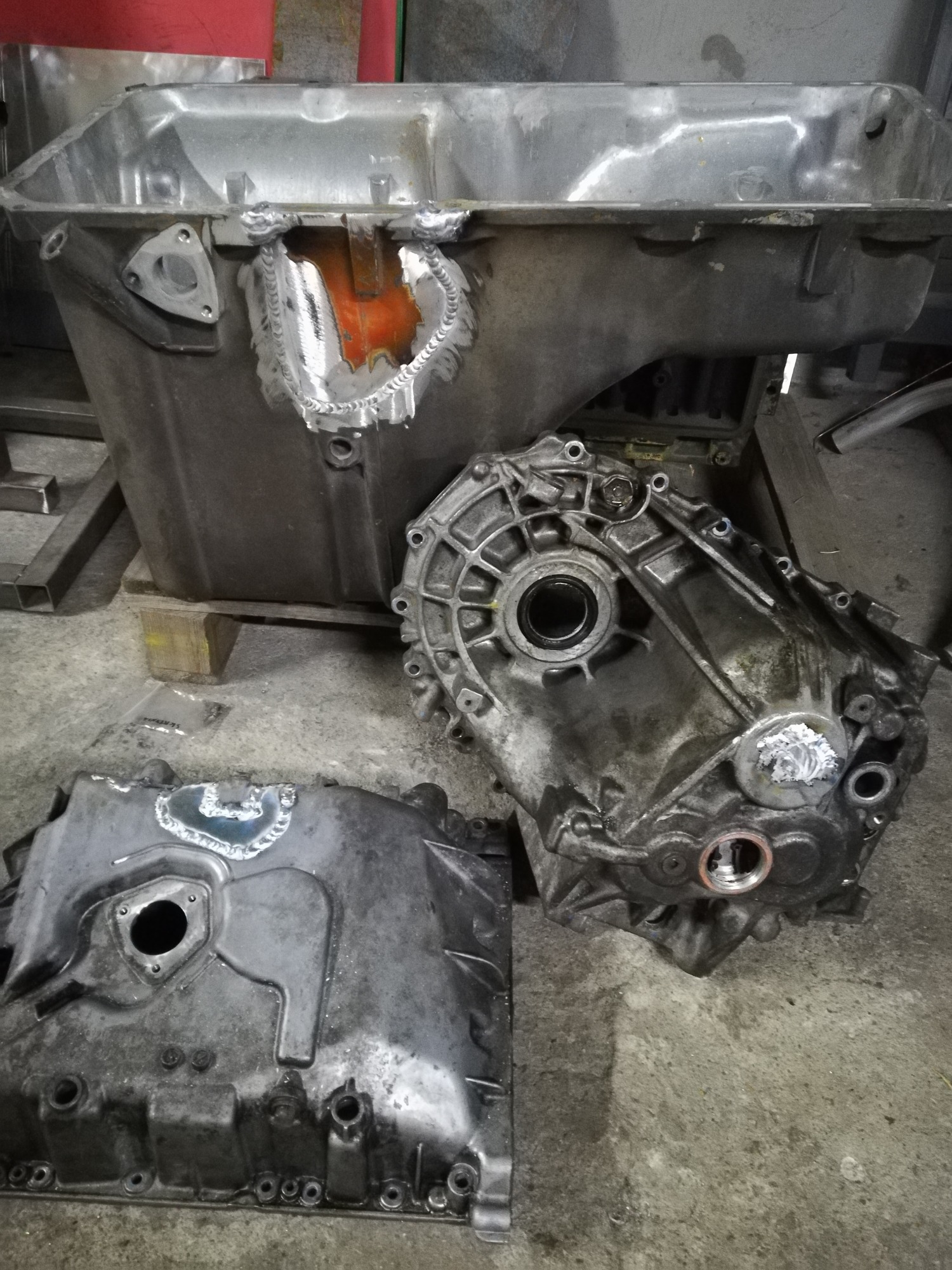Szare, metalowe elementy skrzyni biegów po naprawie, widoczne ślady spawania na obudowie i pokrywie, całość leży na betonowej podłodze.