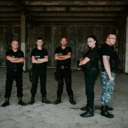 Grupa pięciu ochroniarzy w czarnych uniformach i kamizelkach taktycznych, stojących w rzędzie na tle surowego, betonowego wnętrza z widocznymi śladami graffiti i zniszczeń.