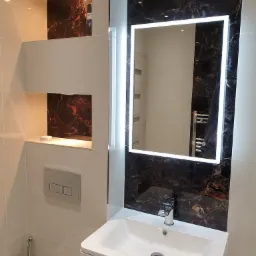 Nowoczesna łazienka z białymi płytkami, umywalką nablatową i podświetlanym lustrem na tle ściany z marmuru. Wnęki z podświetleniem eksponują dekoracyjny marmur. Widoczny fragment toalety z bidetką.