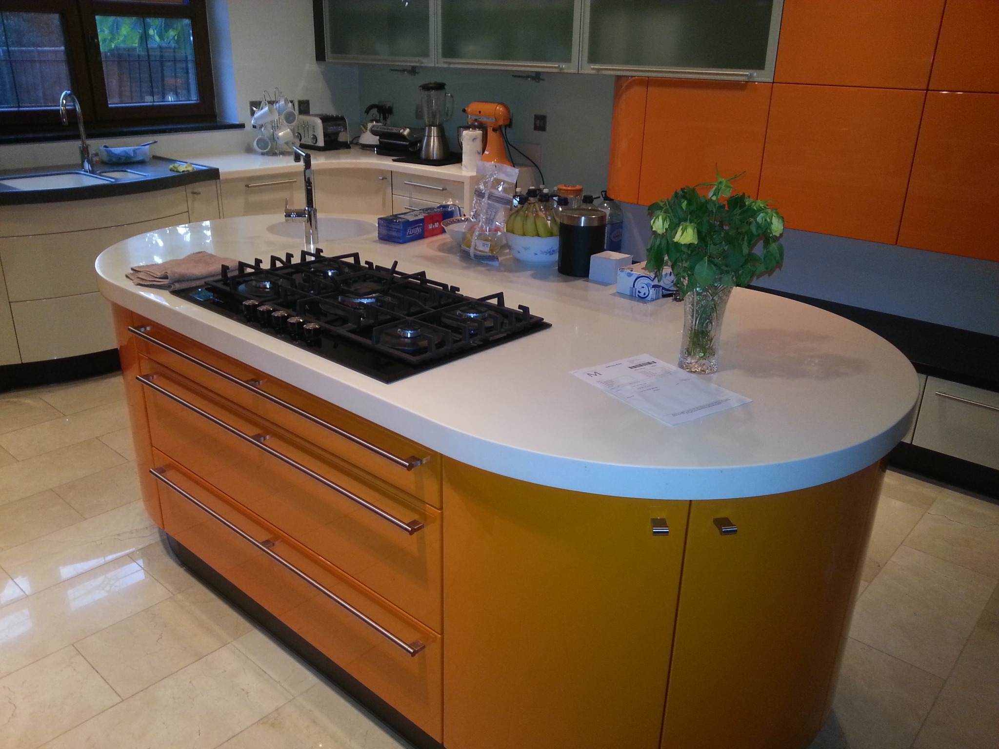 Nowoczesna wyspa kuchenna z pomarańczowymi frontami szafek i białym blatem, na którym znajduje się płyta gazowa, w tle widoczne inne elementy wyposażenia kuchni.