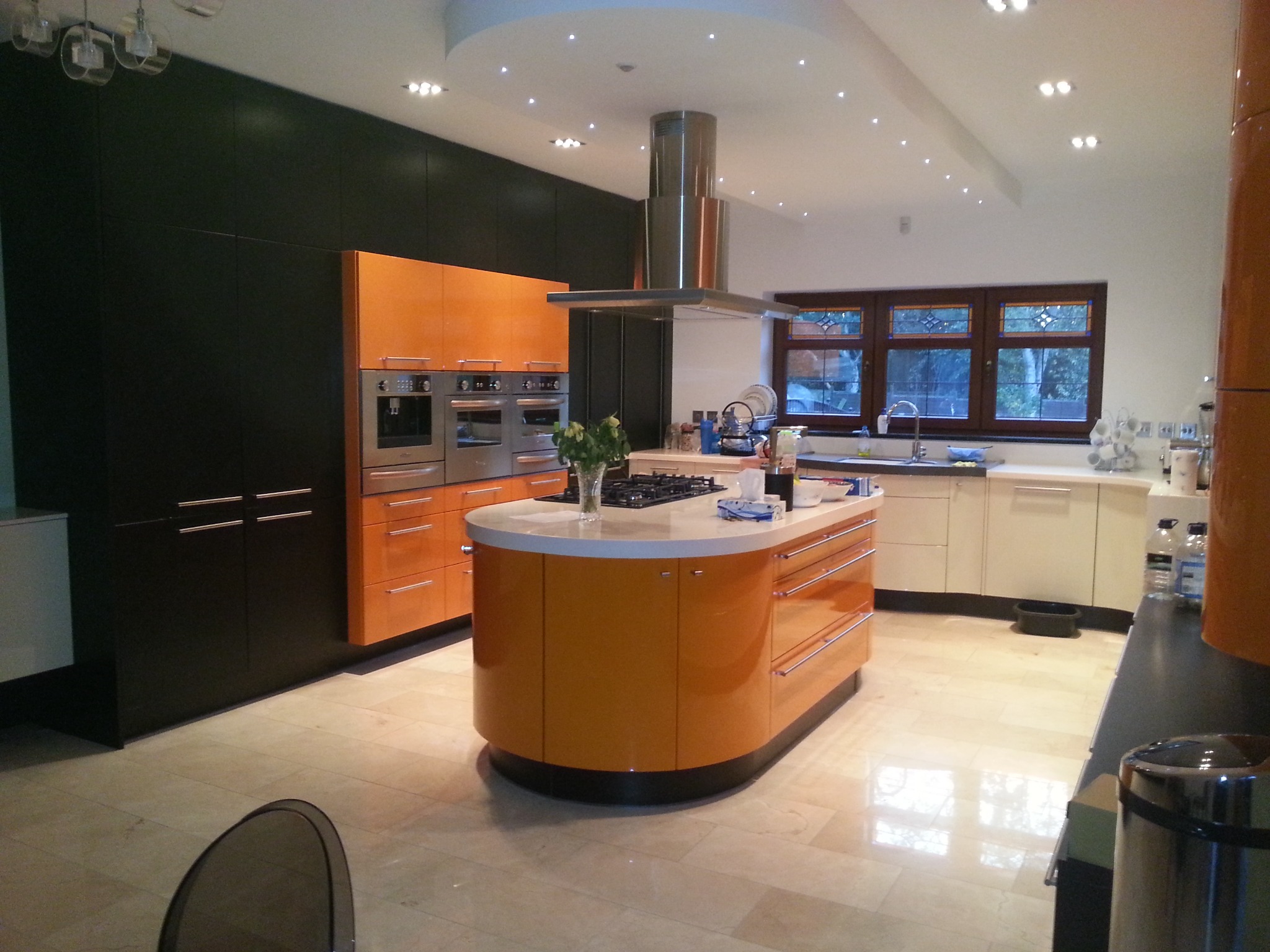Nowoczesna, przestronna kuchnia z wyspą w kształcie elipsy, pomarańczowymi szafkami na wysoki połysk, czarnymi szafami ściennymi i marmurową podłogą. Nad wyspą stalowy okap. Duże okno z drewnianą...