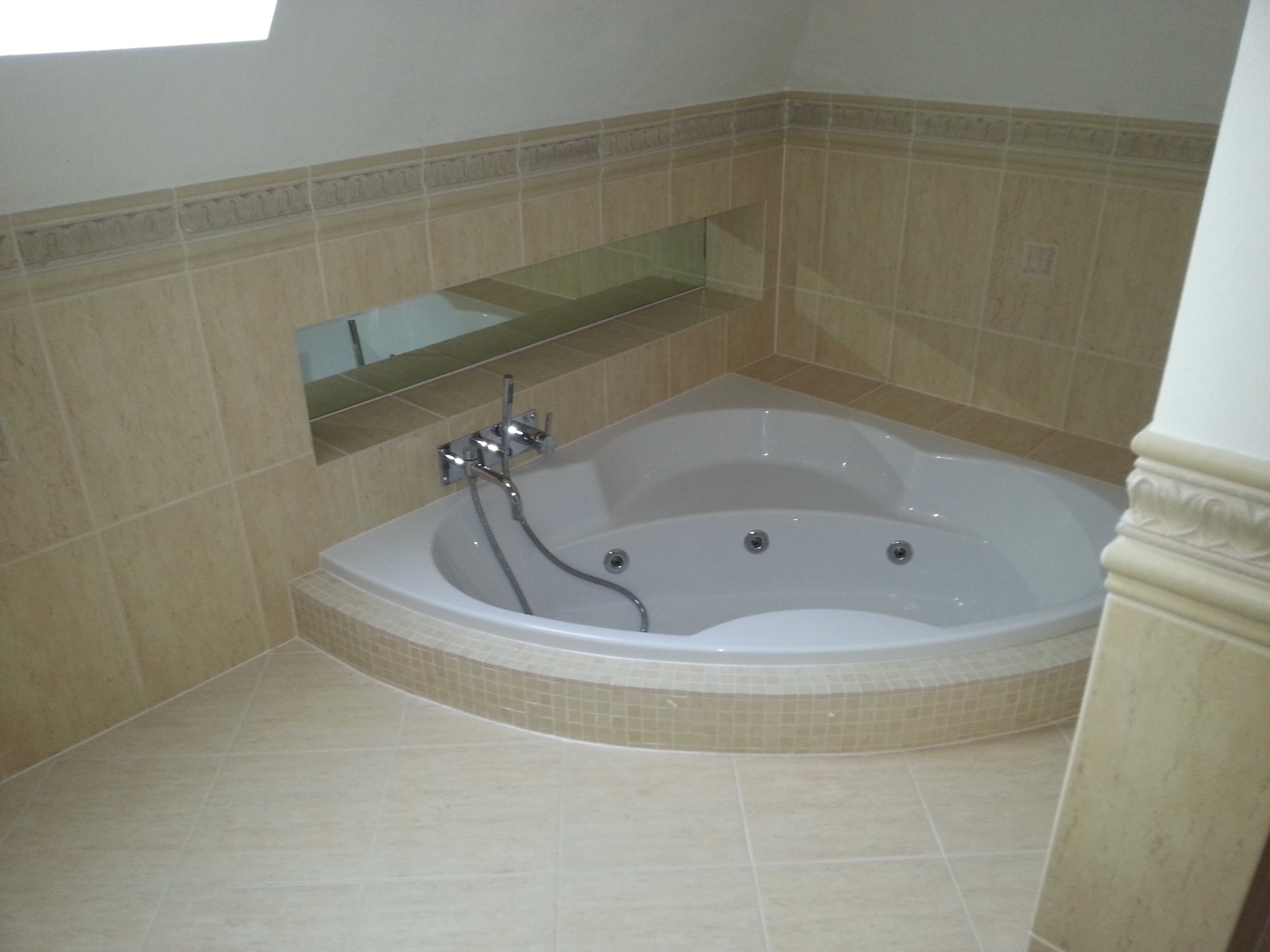 Nowoczesna narożna wanna z hydromasażem, obudowana mozaiką w odcieniach beżu, z chromowaną armaturą i półką w ścianie wyłożoną płytkami, w łazience wykończonej w jasnej tonacji.
