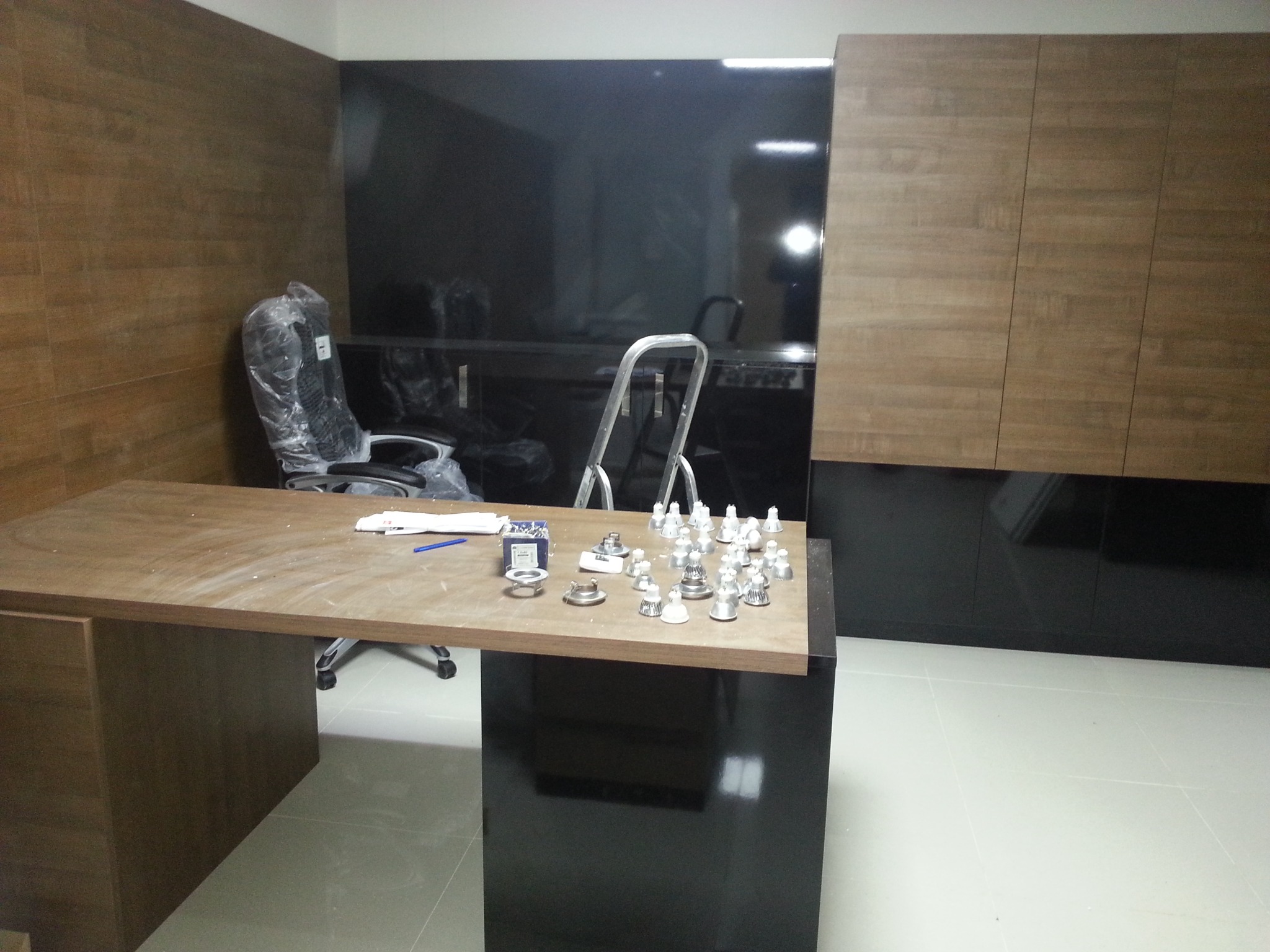 Wnętrze biura przed wykończeniem, widoczny drewniany stół z rozłożonymi elementami oświetlenia, krzesło w folii ochronnej, ciemne ściany i jasna podłoga.