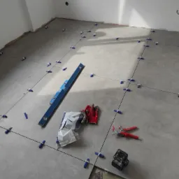 Szare płytki podłogowe w trakcie układania, z użyciem niebieskich klipsów poziomujących, poziomicy, paczki kleju, czerwonych rękawic roboczych i akumulatorowej poziomicy laserowej.