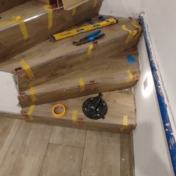 Karmi Bau - Układanie drewnopodobnych płytek na kręconych schodach, z widocznymi klipsami poziomującymi, żółtą taśmą i narzędziami. Częściowo ułożona posadzka na dole, w tle karton z materiałami.