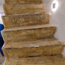 Karmi Bau - Surowe schody pokryte warstwą zaprawy, przygotowywane do wykończenia, z dwoma kwadratowymi lampami ściennymi na białej ścianie. W tle widoczny fragment narzędzia.