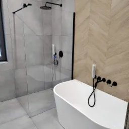 Nowoczesna łazienka z kabiną prysznicową typu walk-in z czarnymi elementami armatury i białą, wolnostojącą wanną na tle ściany z płytek imitujących drewno ułożonych w jodełkę.