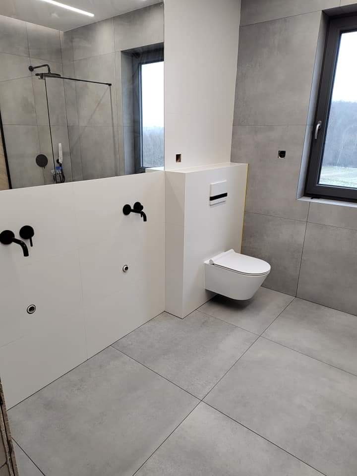 Nowoczesna łazienka z szarymi płytkami, podwieszaną toaletą i czarną armaturą, widok na zewnątrz przez okno.