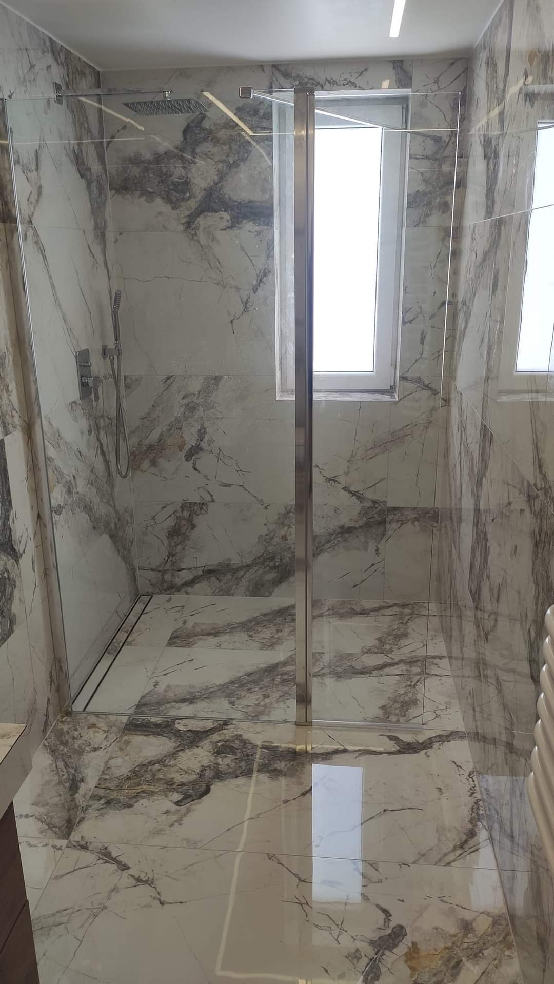 Nowoczesna kabina prysznicowa z marmurowymi płytkami, szklane drzwi z metalowymi okuciami, widoczna deszczownica i odpływ liniowy w podłodze.