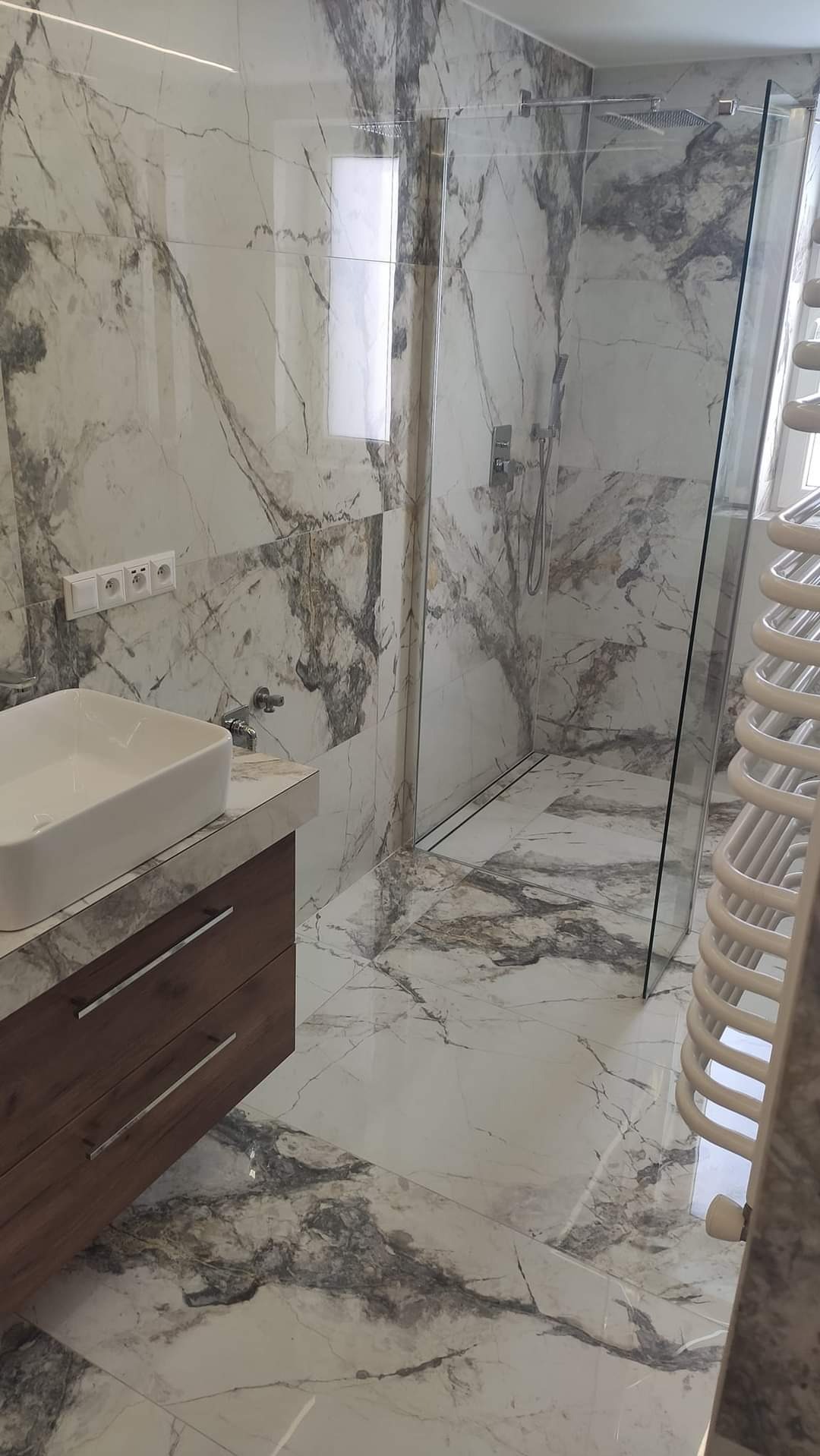 Elegancka łazienka wykończona płytkami imitującymi marmur, z kabiną prysznicową typu walk-in i umywalką nablatową na drewnianej szafce z szufladami.