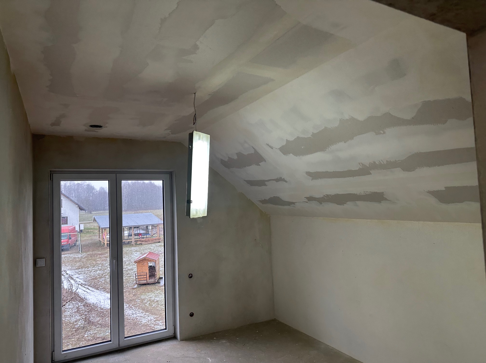 Poddasze w trakcie prac wykończeniowych: ściany i sufit po szpachlowaniu, widoczne nierówności i miejsca po łatach, okno z widokiem na zewnątrz.