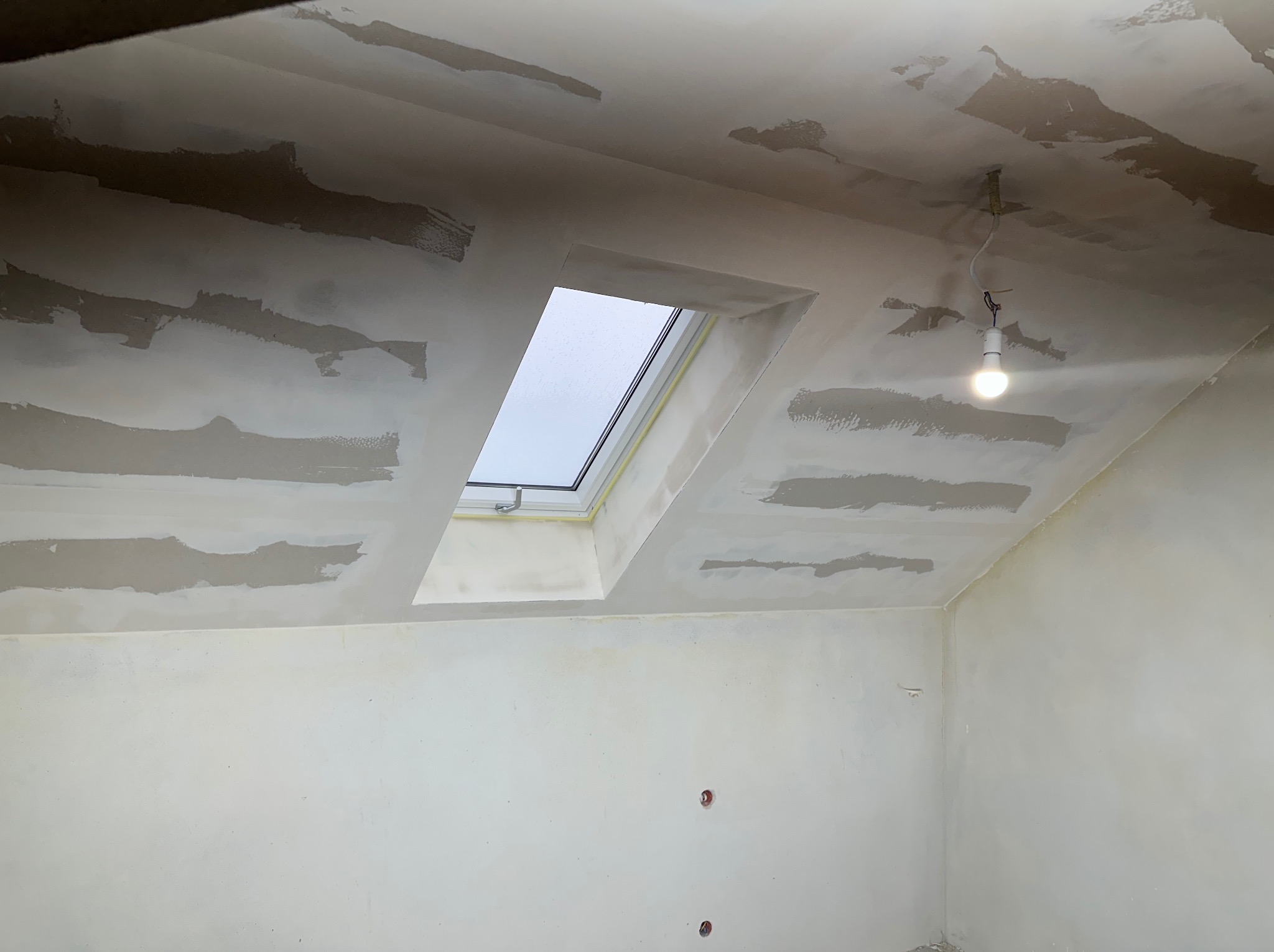 Poddasze w trakcie prac przygotowawczych do malowania, widoczne fragmenty nałożonej gładzi na skośnym suficie oraz zamontowane okno dachowe z żółtą taśmą ochronną, surowa żarówka zwisająca z sufitu.