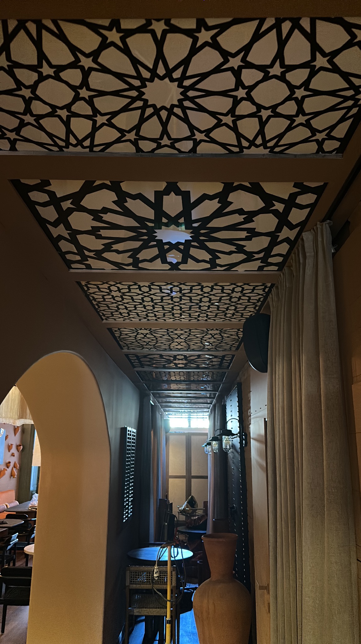 Wnętrze lokalu z dekoracyjnym sufitem w geometryczne wzory, widok w głąb pomieszczenia z elementami wystroju w stylu orientalnym i ciepłą kolorystyką.