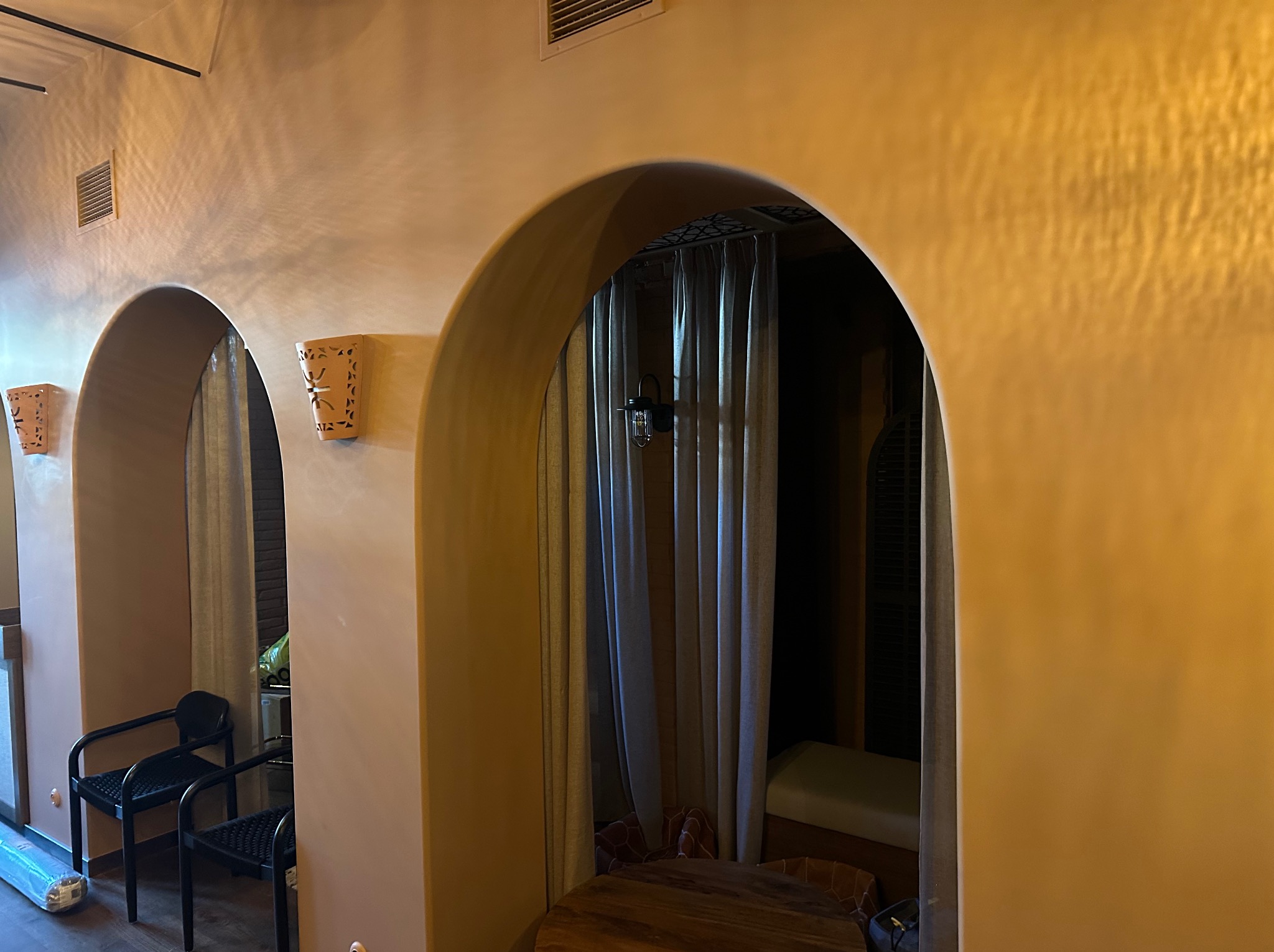 Stylowe wykończenie wnętrza z łukowatymi przejściami i dekoracyjnymi kinkietami. Ściany w ciepłym odcieniu beżu z wyraźną fakturą. Widoczne krzesła i zasłony w tle.