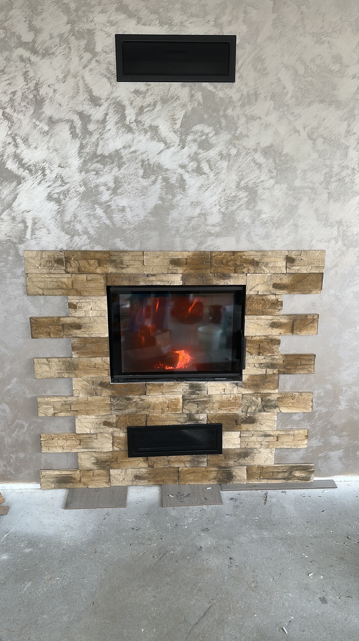 Wykończenie kominka kamieniem dekoracyjnym na ścianie o fakturze betonu. Widoczne elementy montażowe i palenisko z płomieniami. Detal wykończenia wnętrza.