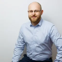 Portret uśmiechniętego mężczyzny z brodą i okularami w błękitnej koszuli na jasnym tle.