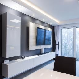 Salon Meblowy WFM Tychy - Nowoczesny salon z szarą ścianą, białymi meblami wiszącymi, telewizorem na ścianie z drewnianym panelem, dużym oknem z białymi zasłonami i czarnymi krzesłami.