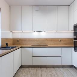 Salon Meblowy WFM Tychy - Nowoczesna kuchnia z białymi szafkami, drewnianym blatem i podłogą z paneli imitujących drewno. Zlewozmywak w kolorze czarnym pod oknem z żaluzjami. Wbudowane sprzęty AGD: piekarnik, mikrofalówka...