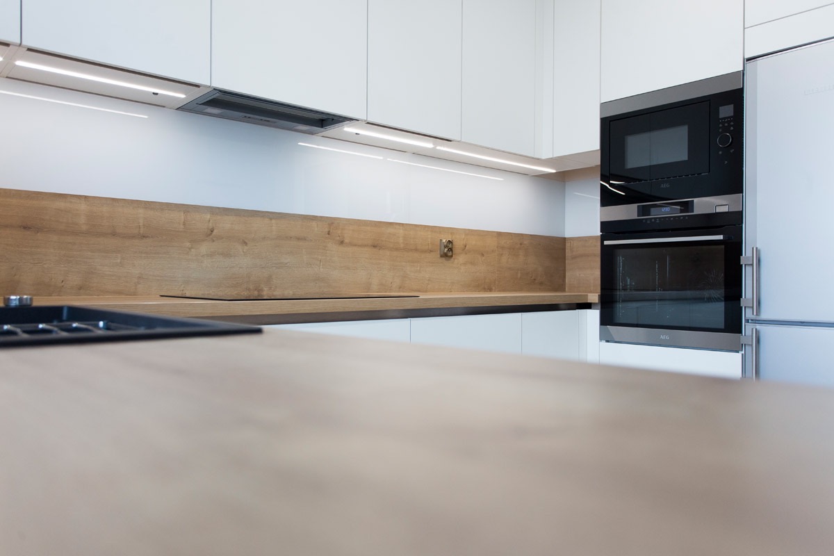 Nowoczesna kuchnia z białymi szafkami, drewnianym blatem i zabudowanym sprzętem AGD marki AEG, oświetlenie LED pod szafkami.