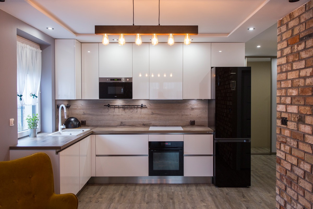 Nowoczesna kuchnia z białymi, lakierowanymi szafkami, drewnianym blatem i podświetleniem LED, z widoczną ścianą z cegły z boku.