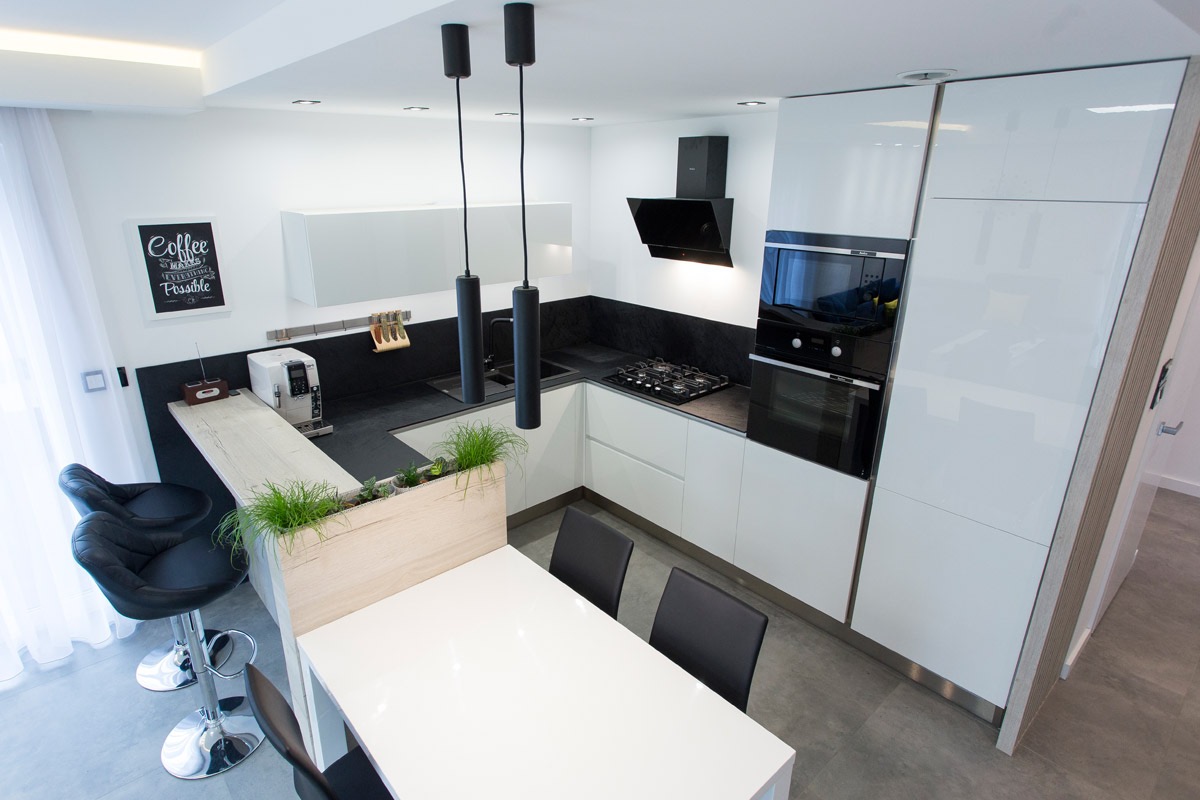 Nowoczesna biało-czarna kuchnia z wyspą oddzielającą przestrzeń roboczą od jadalni, nad którą wiszą dwie czarne lampy cylindryczne. Na blacie stoi ekspres do kawy, a na wyspie donica z zielonymi...