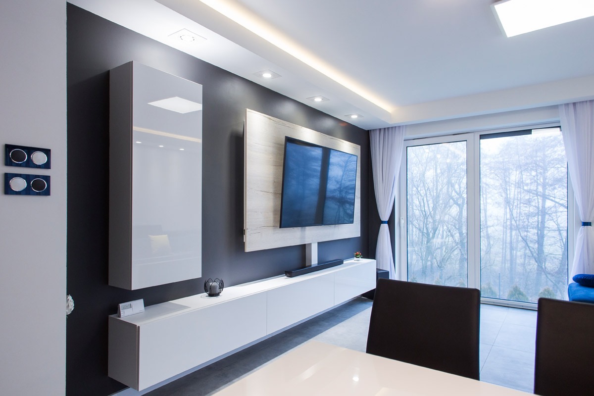 Nowoczesny salon z szarą ścianą, białymi meblami wiszącymi, telewizorem na ścianie z drewnianym panelem, dużym oknem z białymi zasłonami i czarnymi krzesłami.