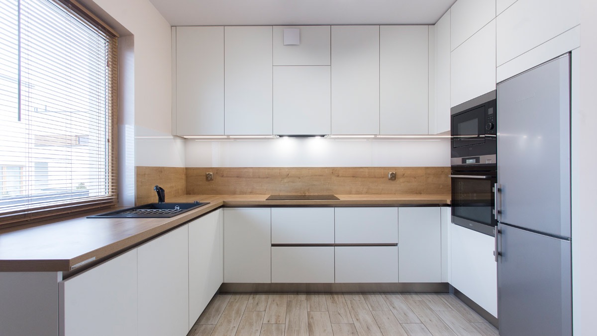 Nowoczesna kuchnia z białymi szafkami, drewnianym blatem i podłogą z paneli imitujących drewno. Zlewozmywak w kolorze czarnym pod oknem z żaluzjami. Wbudowane sprzęty AGD: piekarnik, mikrofalówka...