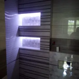 Nowoczesna łazienka z podświetlanymi wnękami wyłożonymi mozaiką trójkątną i płytkami w paski, widoczny fragment instalacji wodno-kanalizacyjnej.