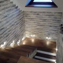 Schody wewnętrzne z drewnianymi stopniami i kamienną okładziną ścian, oświetlone punktowymi lampkami przy podłodze i górnym oknem.