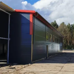 Gotowa hala przemysłowa z szarej płyty warstwowej z czerwonymi elementami wykończeniowymi dachu, widok z zewnątrz w słoneczny dzień.