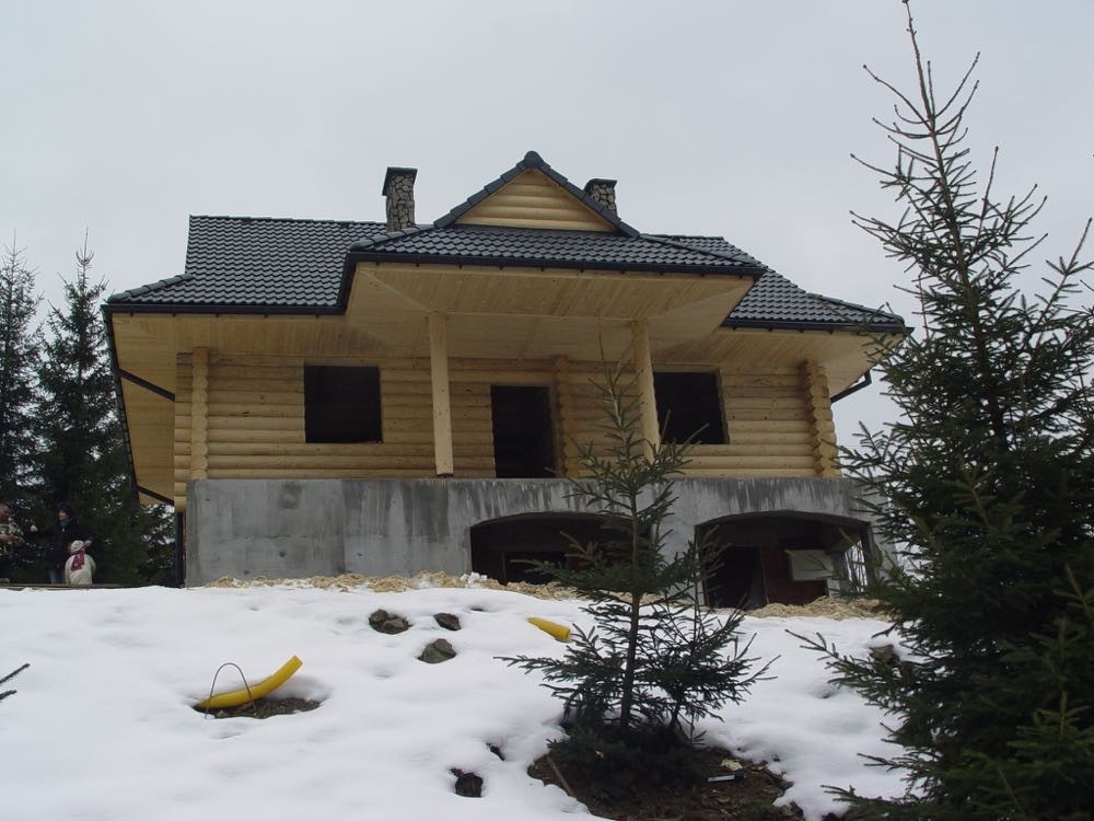 Surowy stan dwukondygnacyjnego domu z bali z ciemną dachówką, widoczny fundament betonowy i zasypany śniegiem teren, na którym stoi bałwan.