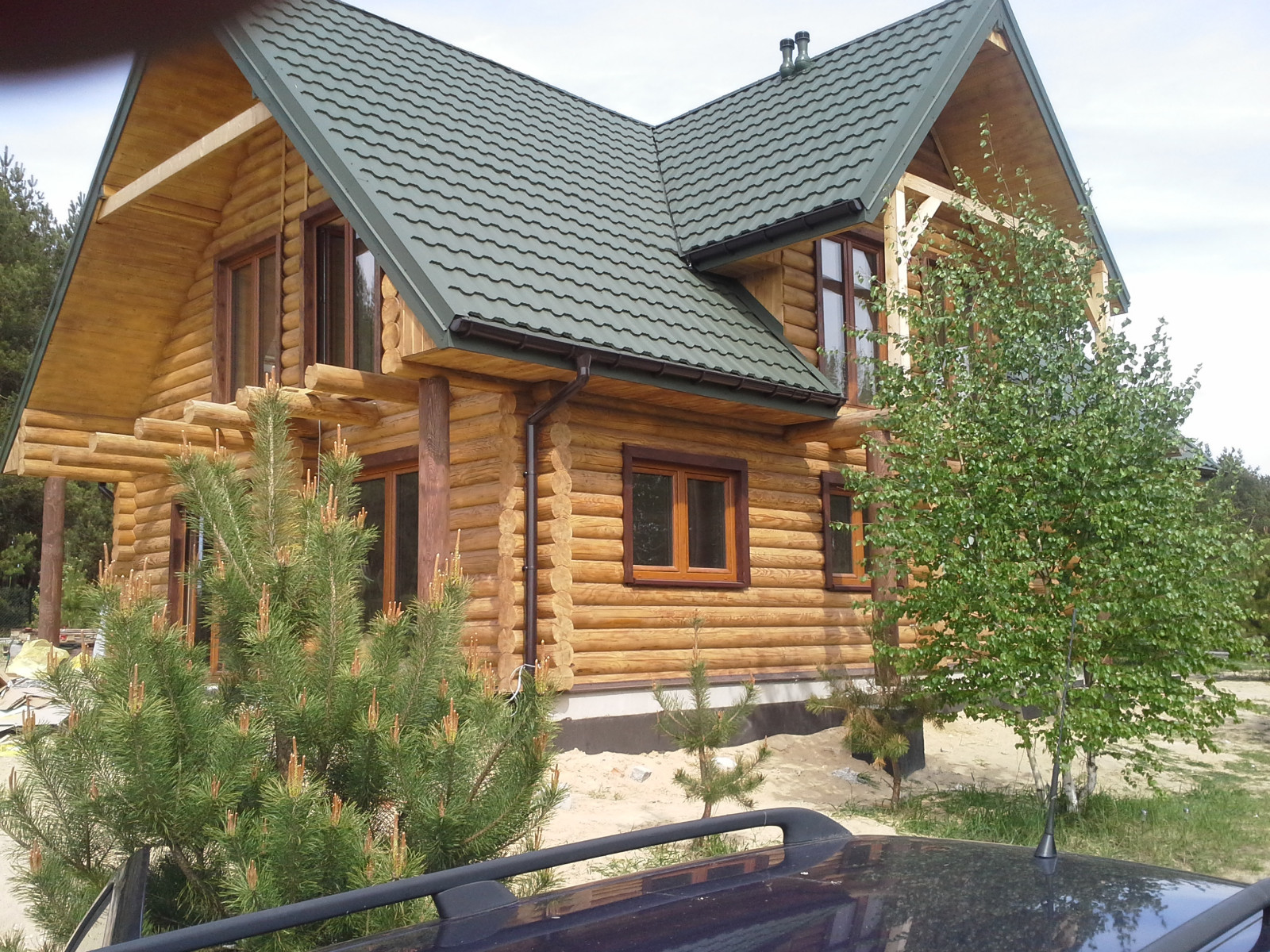 Zewnętrzny widok dwukondygnacyjnego domu z bali z zielonym dachem i brązowymi ramami okiennymi, częściowo zasłoniętego przez drzewa iglaste i brzozę. Widoczny fragment dachu samochodu na pierwszym...