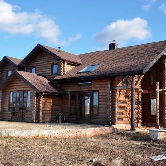 Duży, parterowy dom z bali z brązowym dachem, oświetlony naturalnym światłem słonecznym na tle błękitnego nieba z chmurami.