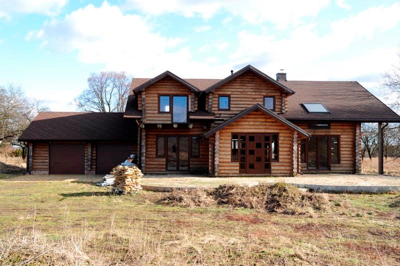 Dwukondygnacyjny dom z bali z brązowym dachem, garażem i stertą kamieni przed budynkiem, uchwycony w słoneczny dzień.