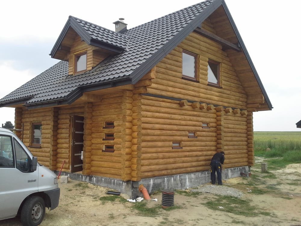 Drewniany dom z bali z ciemnym dachem, częściowo widoczny samochód dostawczy i pracownik wykonujący prace wykończeniowe przy fundamencie.