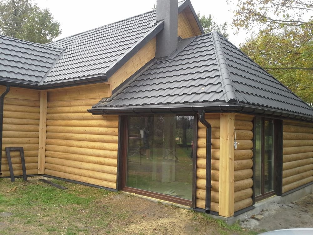 Zewnętrzny widok wykończonego domu z bali z ciemnoszarą dachówką i systemem rynnowym, z widocznymi dużymi oknami z brązowymi ramami.