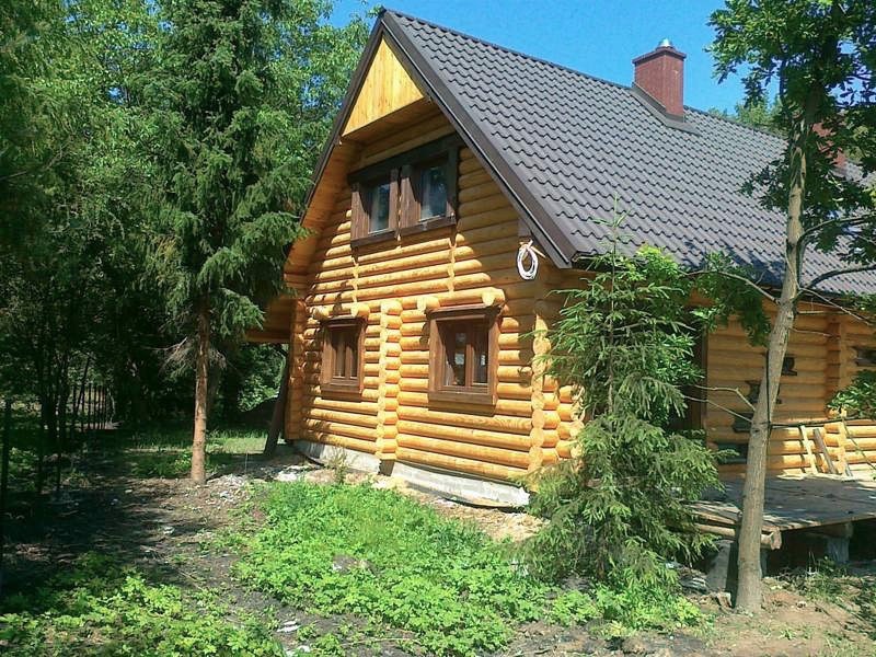 Gotowy dom z bali z ciemnobrązowym dachem, widoczny z zewnątrz, otoczony zielenią drzew i krzewów, z widoczną instalacją elektryczną na elewacji.