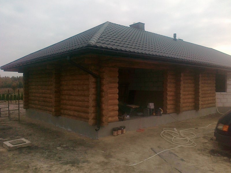 Surowy stan budowy domu z bali z widocznymi puszkami farb i narzędziami wewnątrz, częściowo wykończona elewacja i fragment samochodu w kadrze.