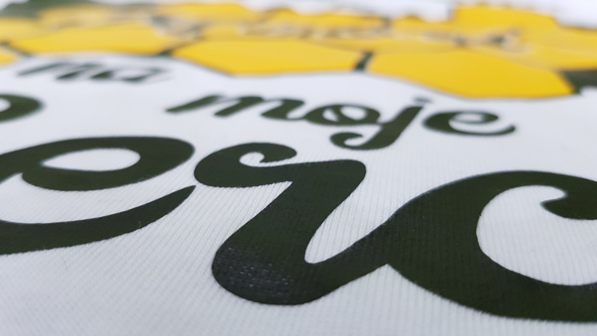 Zbliżenie na białą koszulkę z czarnym, błyszczącym napisem 'na moje serce' i żółtym motywem słonecznika, z efektem głębi ostrości.