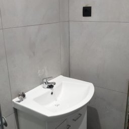 Abecedarian-bud - Biała umywalka narożna z szafką w łazience z szarymi płytkami, widoczny czarny włącznik na ścianie i akcesoria montażowe na umywalce.