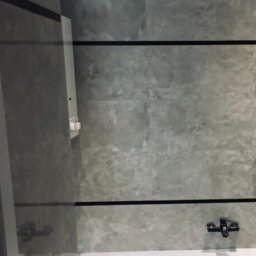 Abecedarian-bud - Wykończona łazienka z szarymi płytkami imitującymi kamień, czarnymi listwami dekoracyjnymi, białą wanną i umywalką z szafką o geometrycznym wzorze.