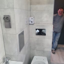 Abecedarian-bud - Wykończona łazienka z szarymi płytkami, białą umywalką i toaletą, widoczny pracownik w tle.