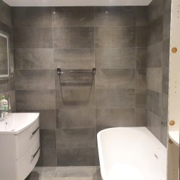 Abecedarian-bud - Wykończona łazienka z szarymi płytkami imitującymi kamień, białą umywalką z szafką i wanną, widoczny fragment ramy drzwiowej z otworami montażowymi.