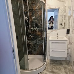 Abecedarian-bud - Wykończona łazienka z kabiną prysznicową z czarnymi płytkami imitującymi marmur, białą umywalką z szafką i lustrem, w którym odbija się osoba robiąca zdjęcie