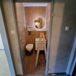 Abecedarian-bud - Remontowana łazienka z drewnopodobnymi płytkami, okrągłym lustrem z podświetleniem LED, toaletą i drabinką malarską w drzwiach; widoczny pojemnik z gruntem Śnieżka.