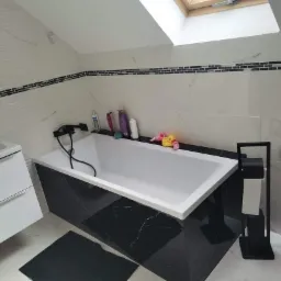 Nowoczesna łazienka na poddaszu z białą armaturą i czarnym akcentem marmuru, widok na wannę z czarną baterią i biało-czarne płytki mozaikowe na ścianach, doświetlona oknem dachowym.