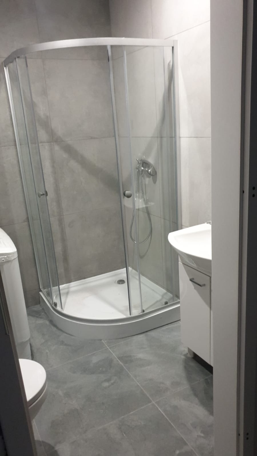 Nowoczesna łazienka z narożną kabiną prysznicową z transparentnego szkła i srebrnymi profilami, umywalką na szafce i częściowo widoczną toaletą, wykończona szarą płytką wielkoformatową.