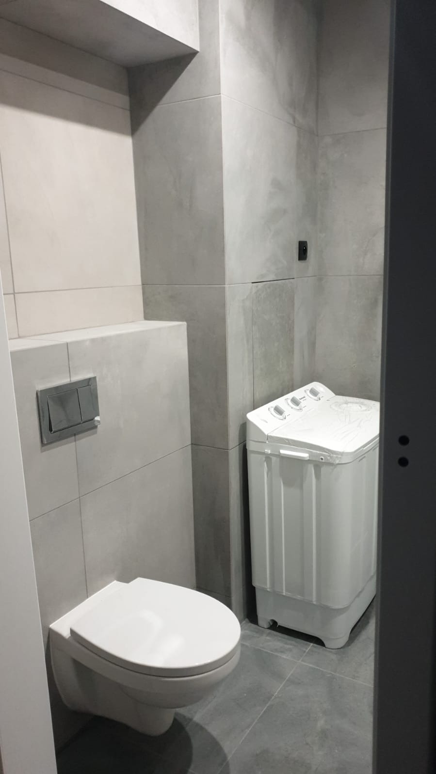 Szara łazienka z wiszącą toaletą, podtynkowym systemem spłukiwania i małą, białą pralką automatyczną.